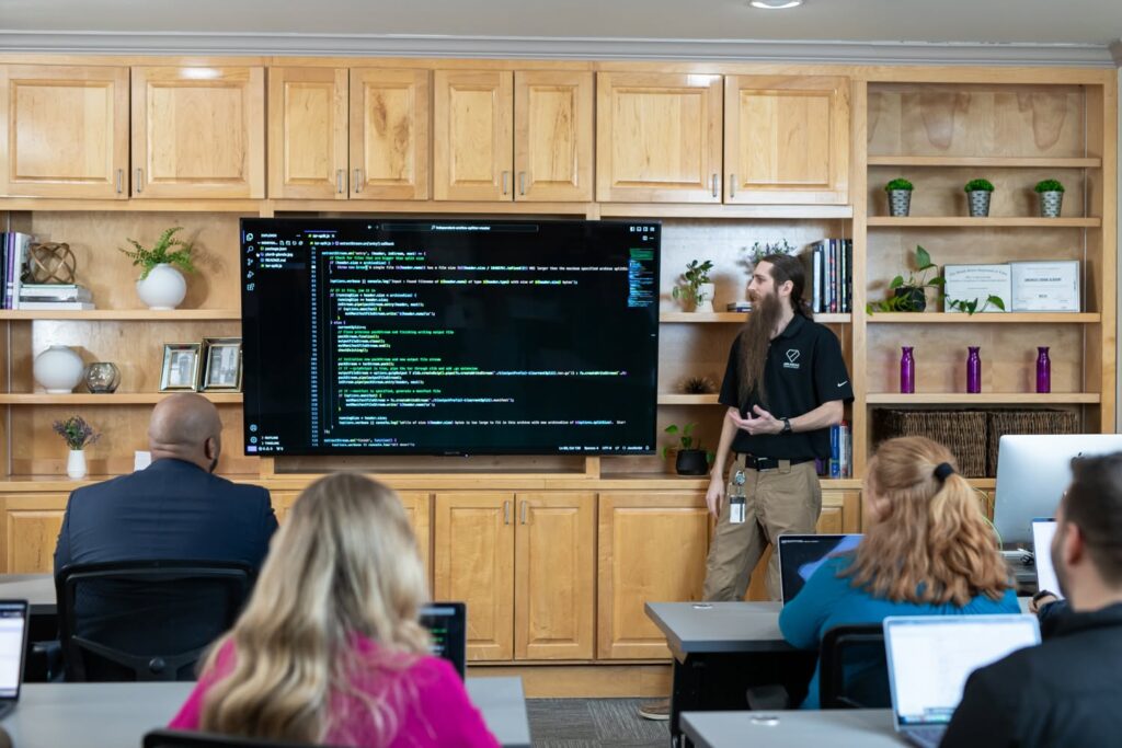 In a classroom with students, an instructor stands next to a large TV showing lines of code.